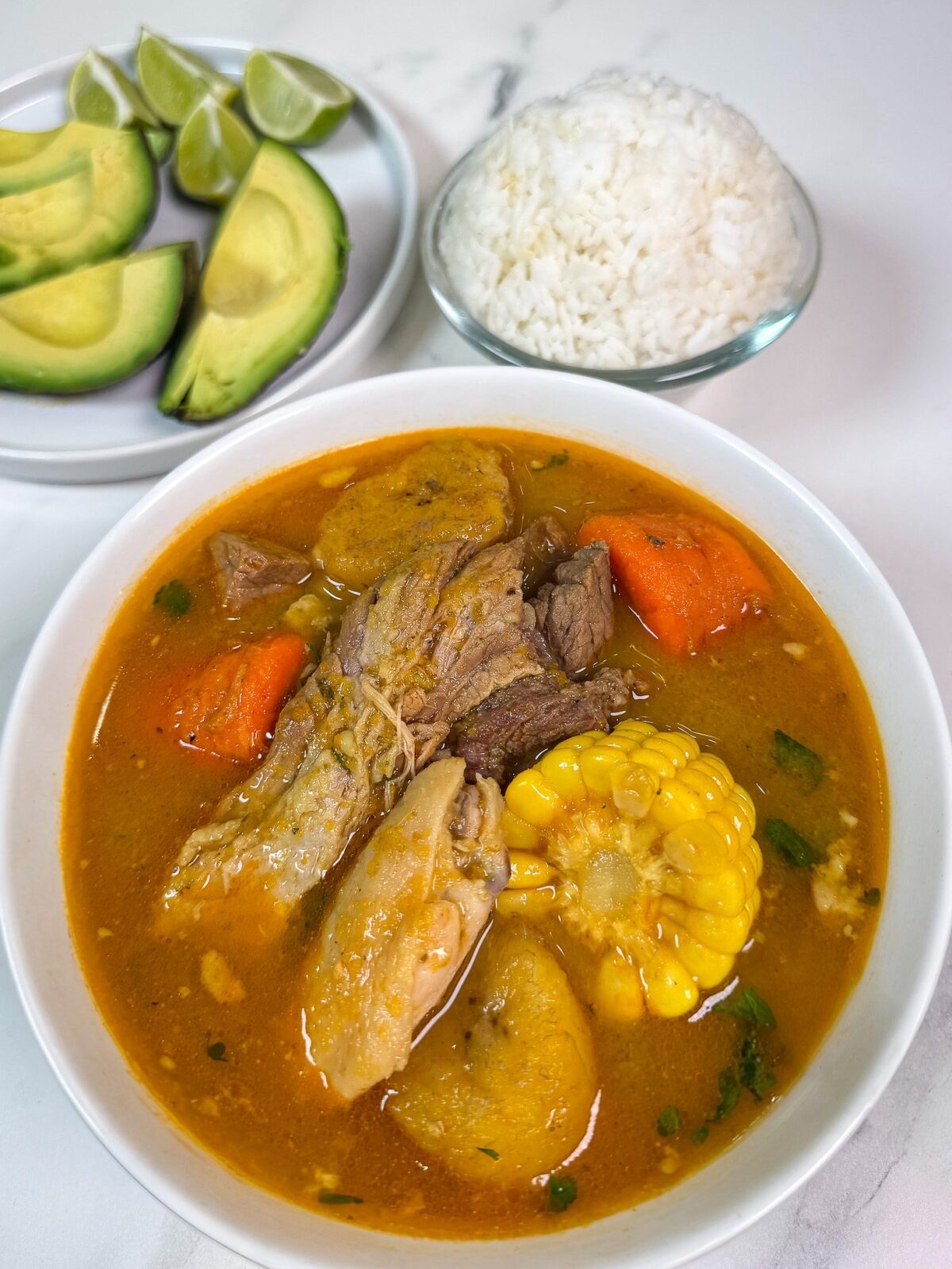 Bowl of Puerto Rican sancocho with beef, chicken, corn, carrots and plantains, served with white rice, avocado and lime