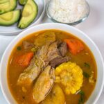 Bowl of Puerto Rican sancocho with beef, chicken, corn, carrots and plantains, served with white rice, avocado and lime