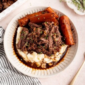 A plate of red wine braised pot roast with whole carrots and mashed potatoes on a striped linen napkin