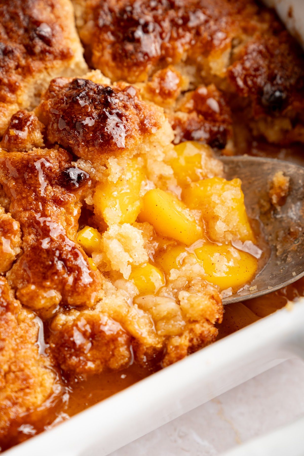 A spoon lifting a portion of biscuit top peach cobbler out of the pan with the peach filling bubbling underneath
