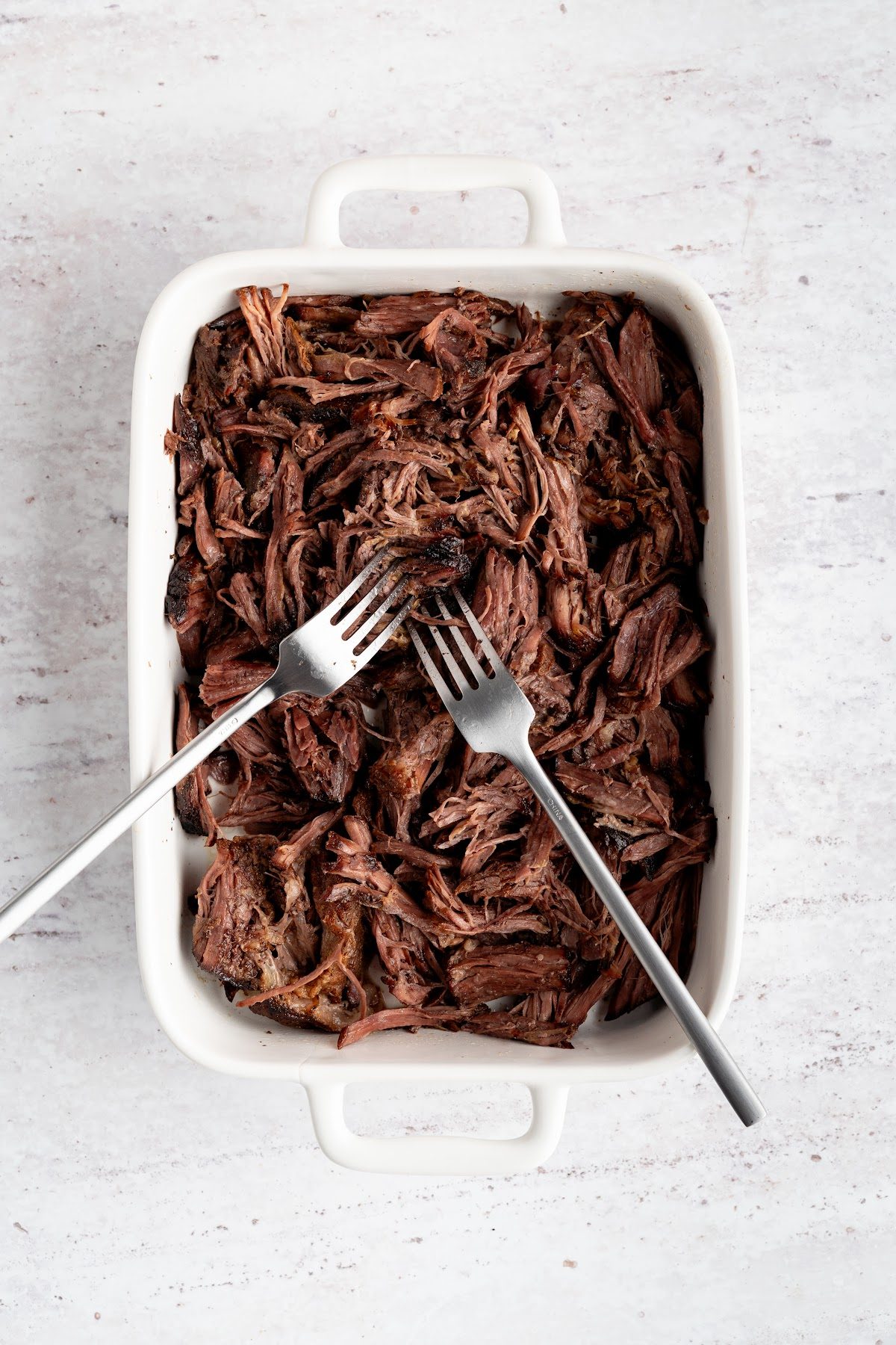 Two forks shredding the braised chuck roast in a white baking dish