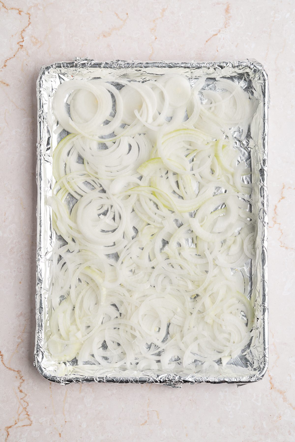 Thinly sliced onions spread in an even layer across a foil-lined half sheet pan