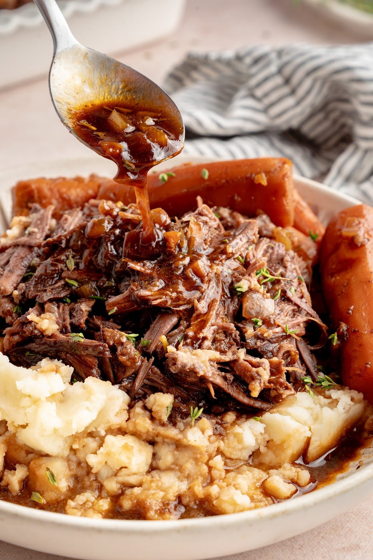A spoon pouring dark red wine gravy over shredded pot roast and mashed potatoes