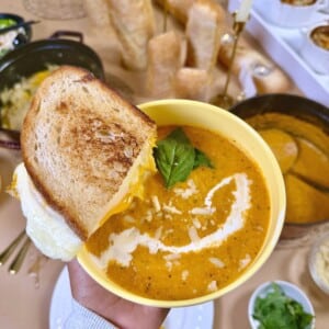 A garlic grilled cheese held over a bowl of roasted tomato soup topped with basil, cream and Parmesan.