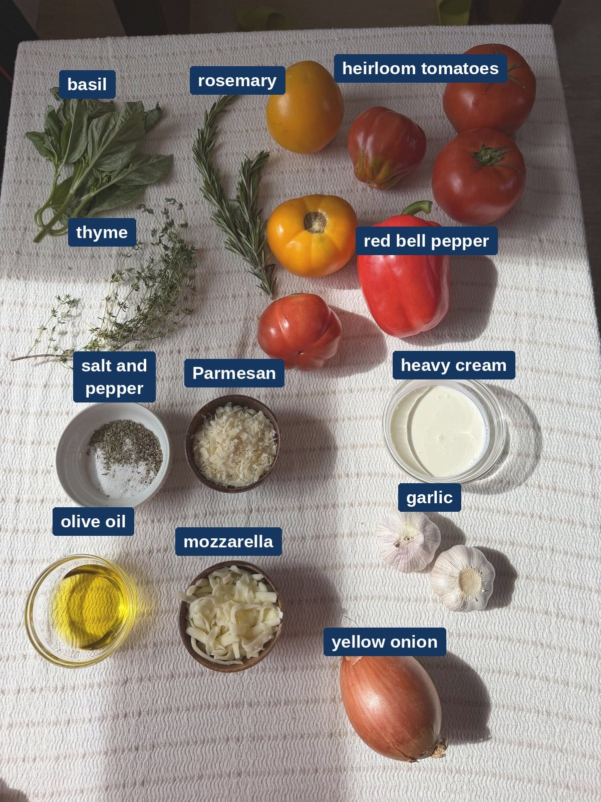 All of the ingredients for roasted tomato soup laid out on a counter, including heirloom tomatoes, red bell pepper, garlic and fresh herbs.