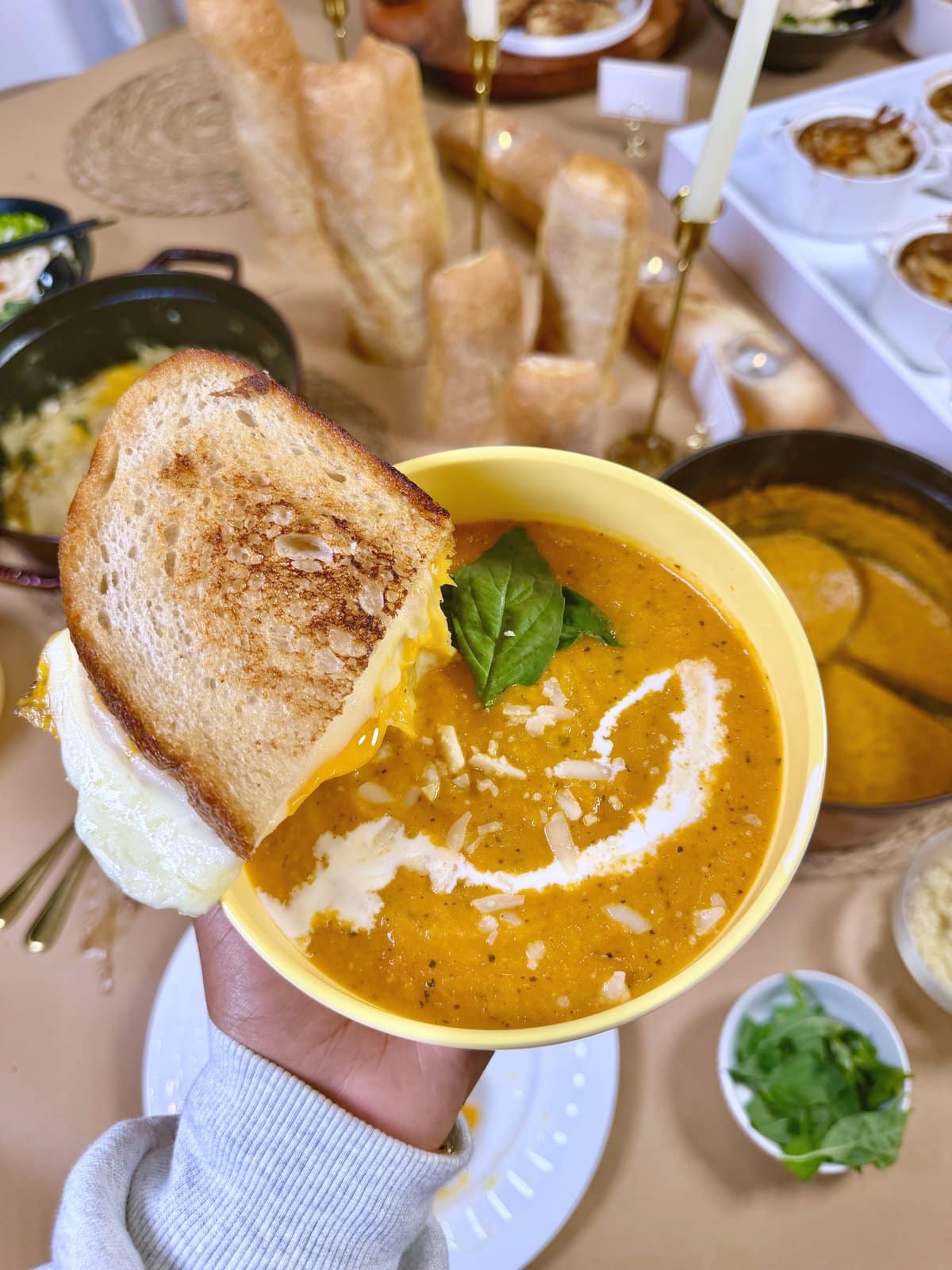 A garlic grilled cheese held over a bowl of roasted tomato soup topped with basil, cream and Parmesan.